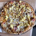 A Breakfast Pizza Topped With Scrambled Eggs, Cheese, And Ham On A Wooden Table., Bevy's Tavern,